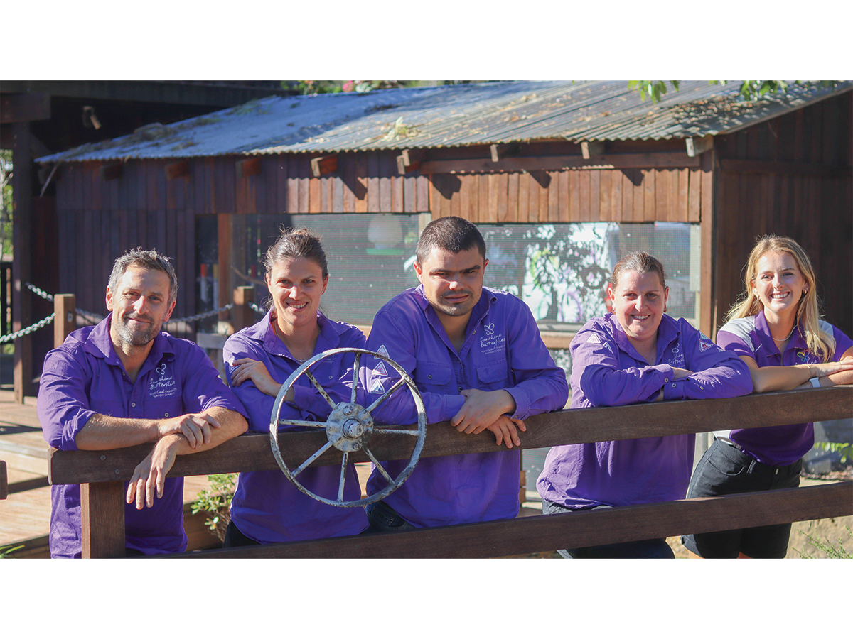 A group of people, some with visible disabilities, dressed in purple uniforms and leaning on a fence outside a rustic looking shed.
