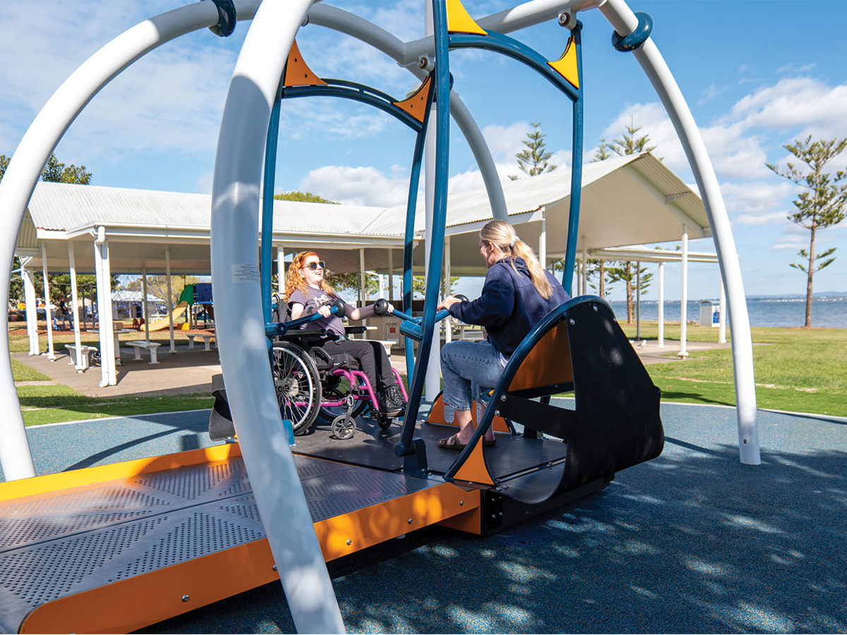 A large wheelchair accessible swing at a beachside park in the sun, with two people using the swing, one in a wheelchair.
