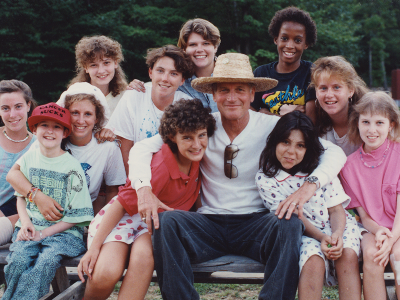 A middle aged man in a straw hat and white shirt and dark pants sits with a group of kids in colourful clothes in a casual photo from the 1990s.