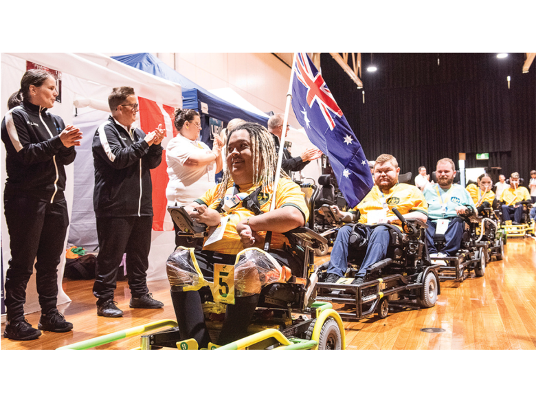 A line of athletes in power chairs, rolling past people applauding them from the side of the image.