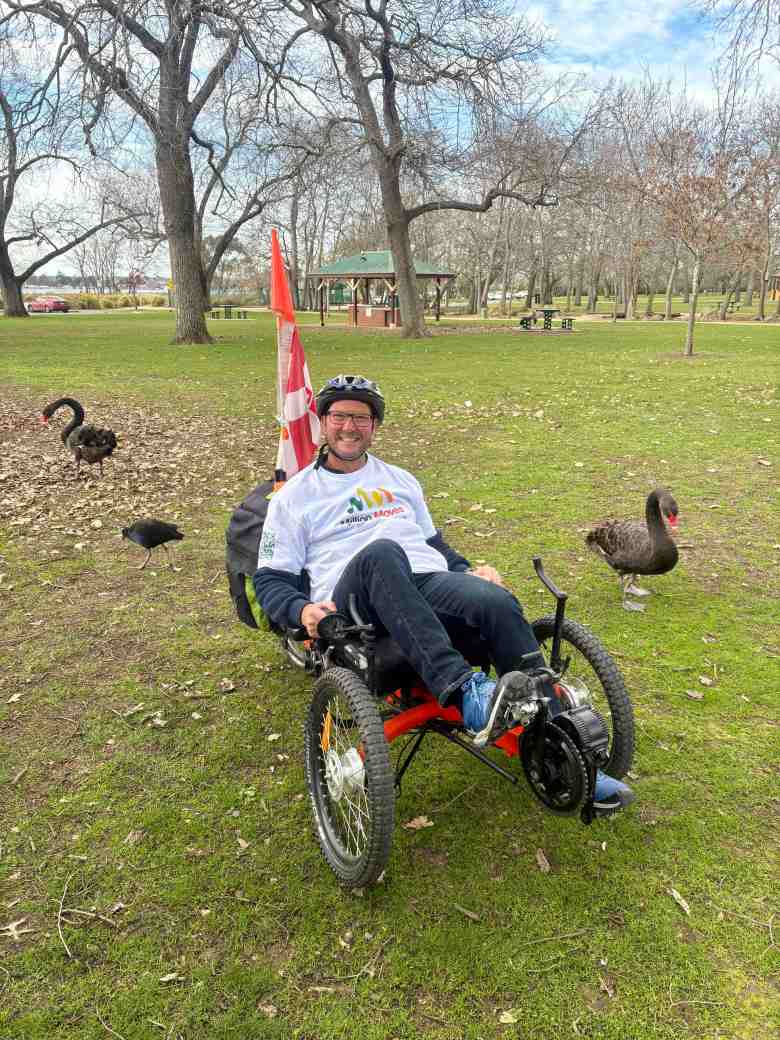 Evan Milne, a middle aged man in a white shirt and black pants, on a trike in a field surrounded by birds. He is part of Million Moves.
