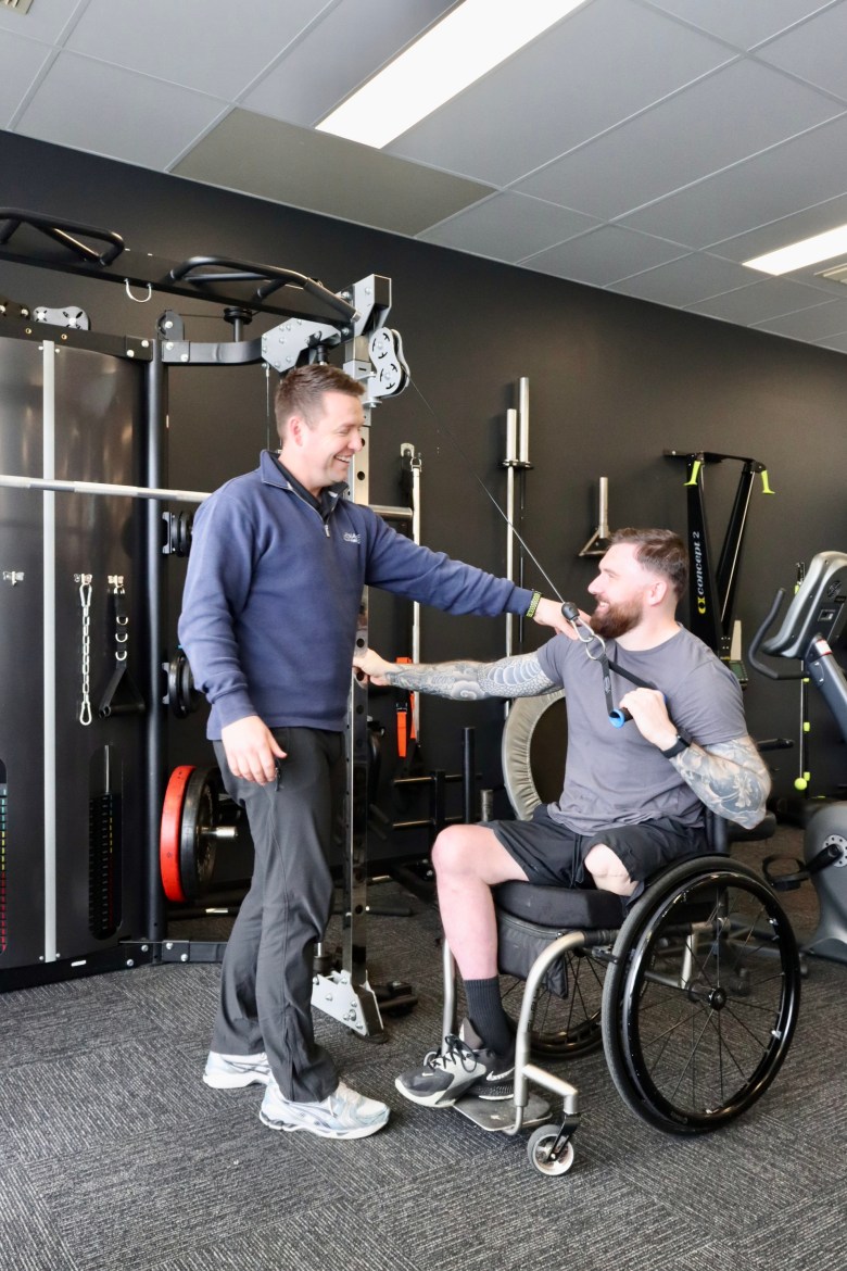 Two men, including one in a wheelchair and one standing, in front of gym equipment.