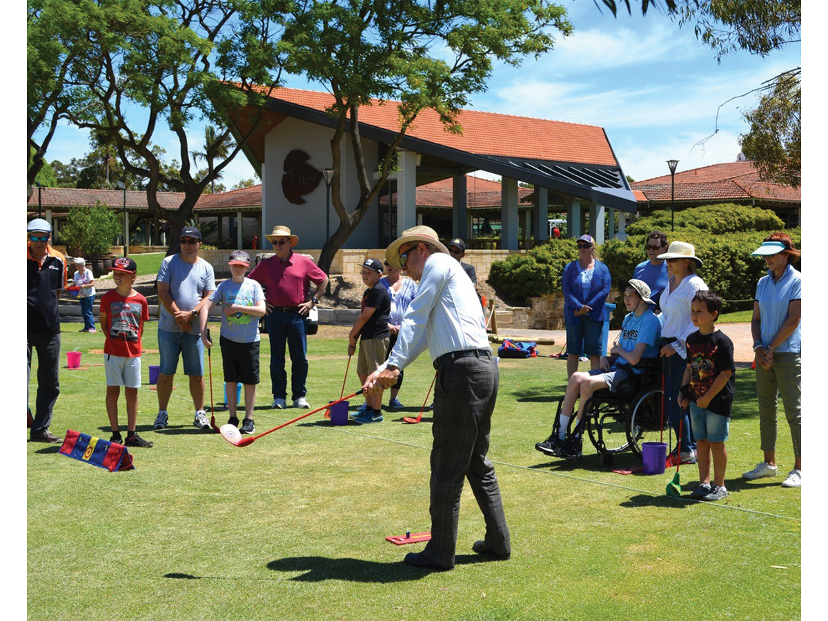 A man outside is swinging a golf club in front of a crowd of people with disability, on a sunny day.