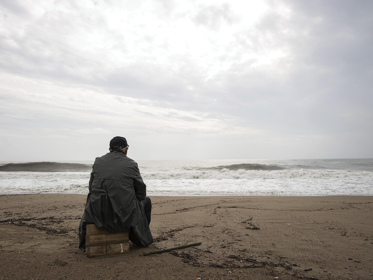 A lonely man wrapped in a jacket, sitting on a gloomy beach looking out to the water. We see him from behind.
