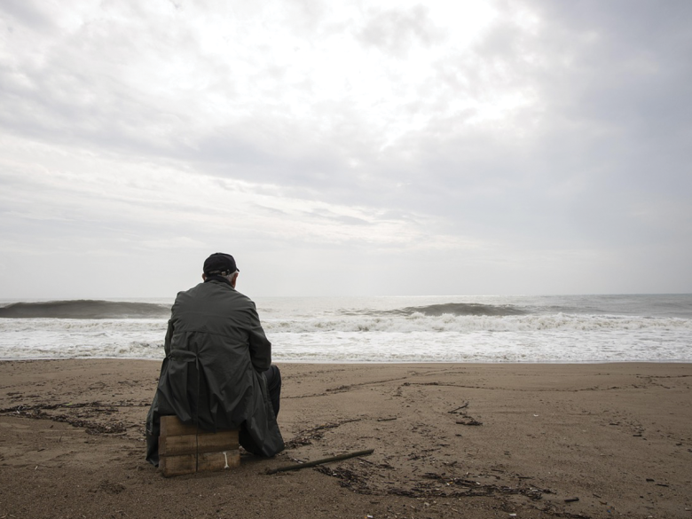 A lonely man wrapped in a jacket, sitting on a gloomy beach looking out to the water. We see him from behind.