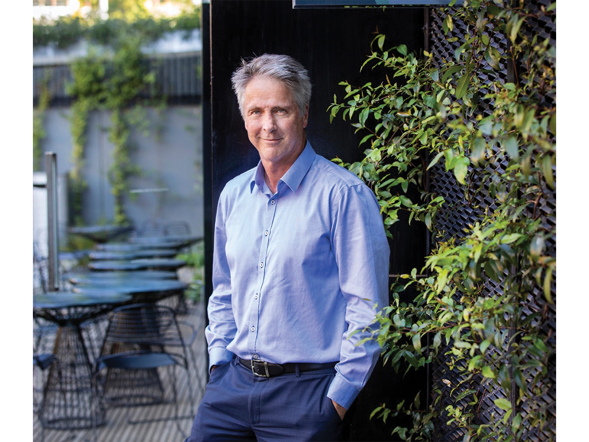 A middle aged man with grey hair in a blue business shirt , stands in a garden environment with his hands in his pockets.