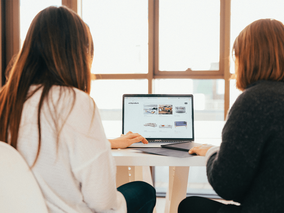 Two women sitting facing a computer, looking at something on the screen together. The background office is light and bright.