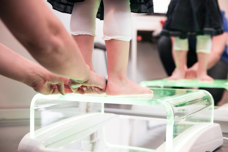 A child's feet on a perspex stand with an adult hand behind fixing their stance. We only see from the feet level.