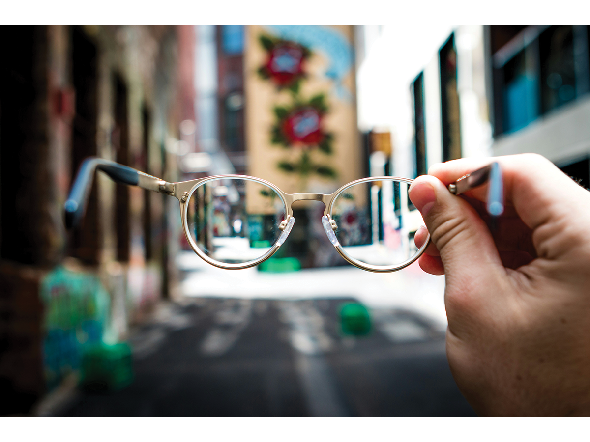 A hand holds up a pair of glasses, we can see a pretty streetscape through them but it is blurry.