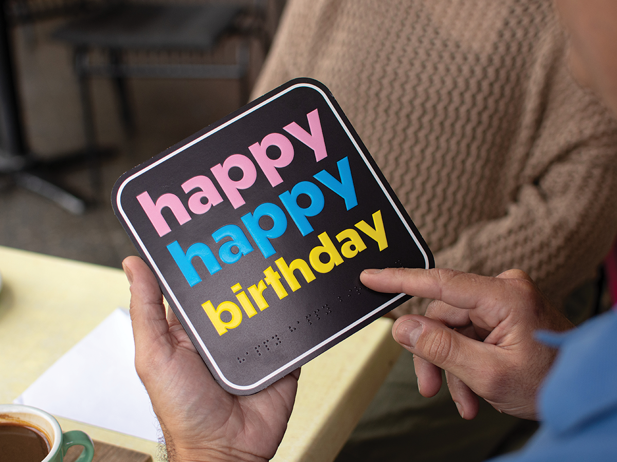 We see a pair of hands holding a birthday card that says Happy Happy Birthday in colourful writing with braille underneath.