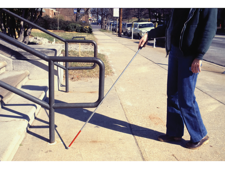 We see a person from the waist down holding a white cane, standing in front of a flight of stairs.
