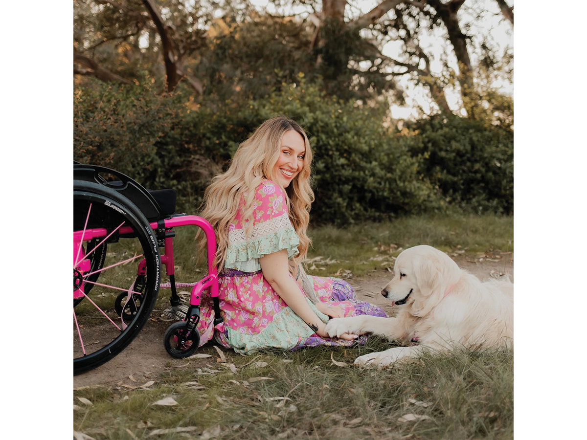 Rhiannon Tracey, a young blonde woman in a colourful dress, sits in a garden next to a pink wheelchair and a golden retriever dog.