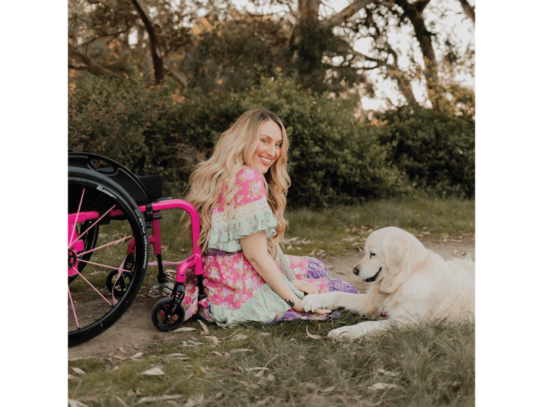 Rhiannon Tracey, a young blonde woman in a colourful dress, sits in a garden next to a pink wheelchair and a golden retriever dog.