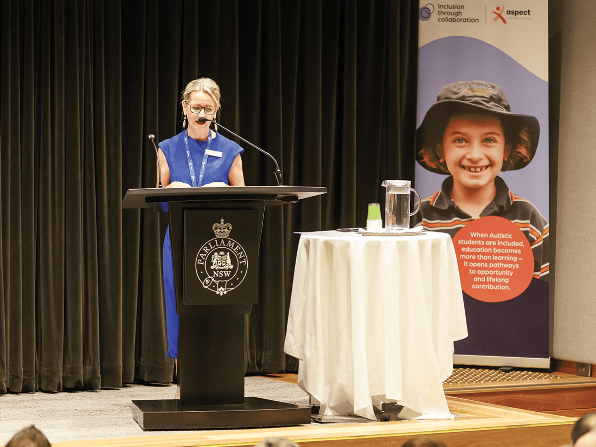 A lady in a blue dress, with blonde hair pulled back, stands behind a lectern on a stage, looking down at notes. There is a small round table with a white tablecloth next to her and a banner behind her with a smiling child on it.