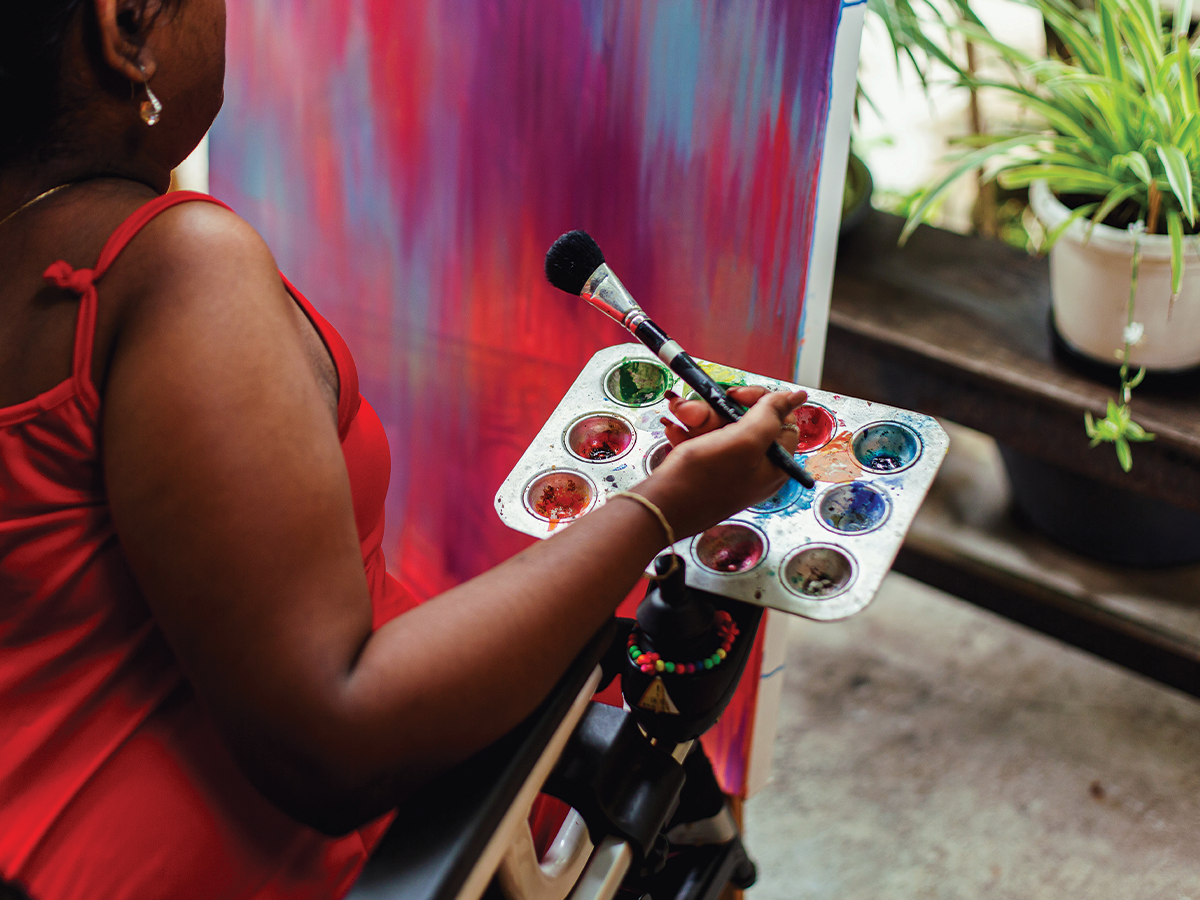 A young black women in a wheelchair holds a colourful paint palette on her knee. We can only see her side. There is a bright pink and blue canvas in front of her.